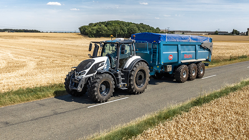A large, modern black Valtra tractor is captured driving down a paved rural road. The vehicle dominates the foreground, with expansive countryside and cloudy skies in the background. The tractor's robust tires and mechanical details are clearly visible, emphasizing its power and utility. No visible text, numbers, or identifiable brands are present in the image.