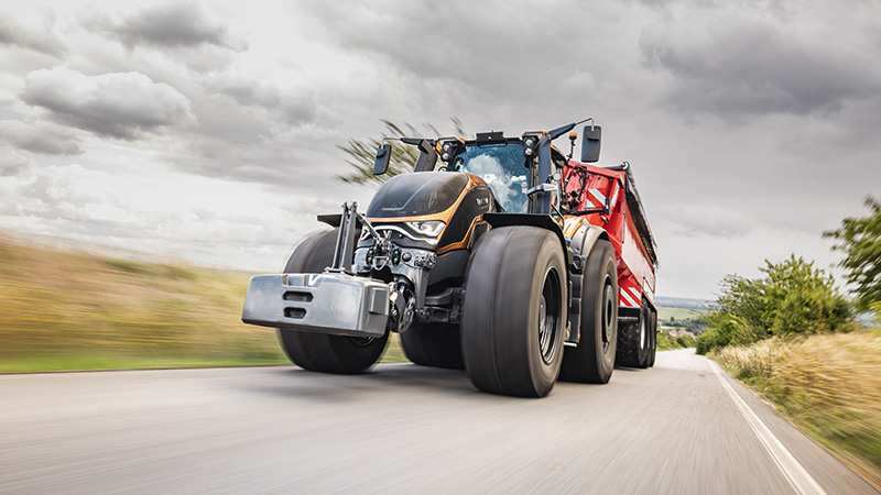 A large, modern S Series Valtra tractor is captured in motion, hauling a red trailer along a paved rural road. The scene is set under a dramatic, cloudy sky, with blurred grass and fields indicating speed. The tractor's robust design and oversized tires are prominent, emphasizing power and utility. No visible text, numbers, or identifiable brands are present in the image.